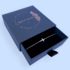 Anklet in silver with a stunning gem encrusted cross within the chain, presented in a blue jewellery box with Anklet Allure written on the top in rose gold.