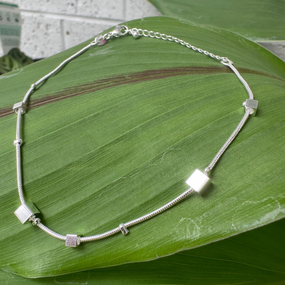 B2-Creativity-Anklet Shiny silver anklet with five silver square charms around it presented on a big green leaf.