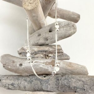 Double chain silver anklet with a heart between two silver balls, repeated three times around the chain, hanging from white washed wood pieces piled up.