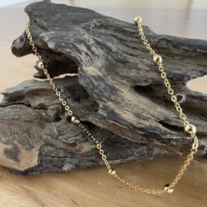 Elegant gold anklet with small gold balls spread around a gold chain at equal distance. Displayed on a piece of driftwood.