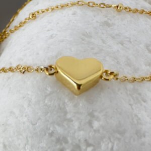 Close up of a dainty gold double chain anklet with a sold gold heart charm on a white rock.