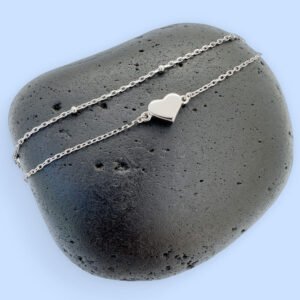 Double chained silver anklet with a solid silver heart charm displayed on a black rock with a blue background.