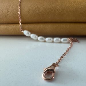 Rose gold ankle chain with the clasp in the foreground and six freshwater pearls in a line in the background draped over a mustard coloured bag.