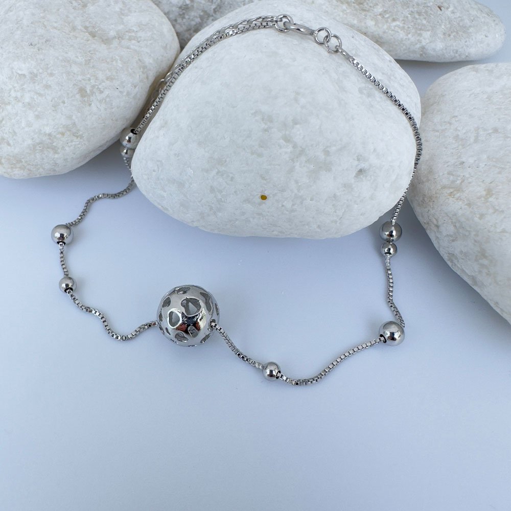 D8-Anklet-(1) Silver anklet with a sphere charm and smaller balls around the chain draped over white rocks.