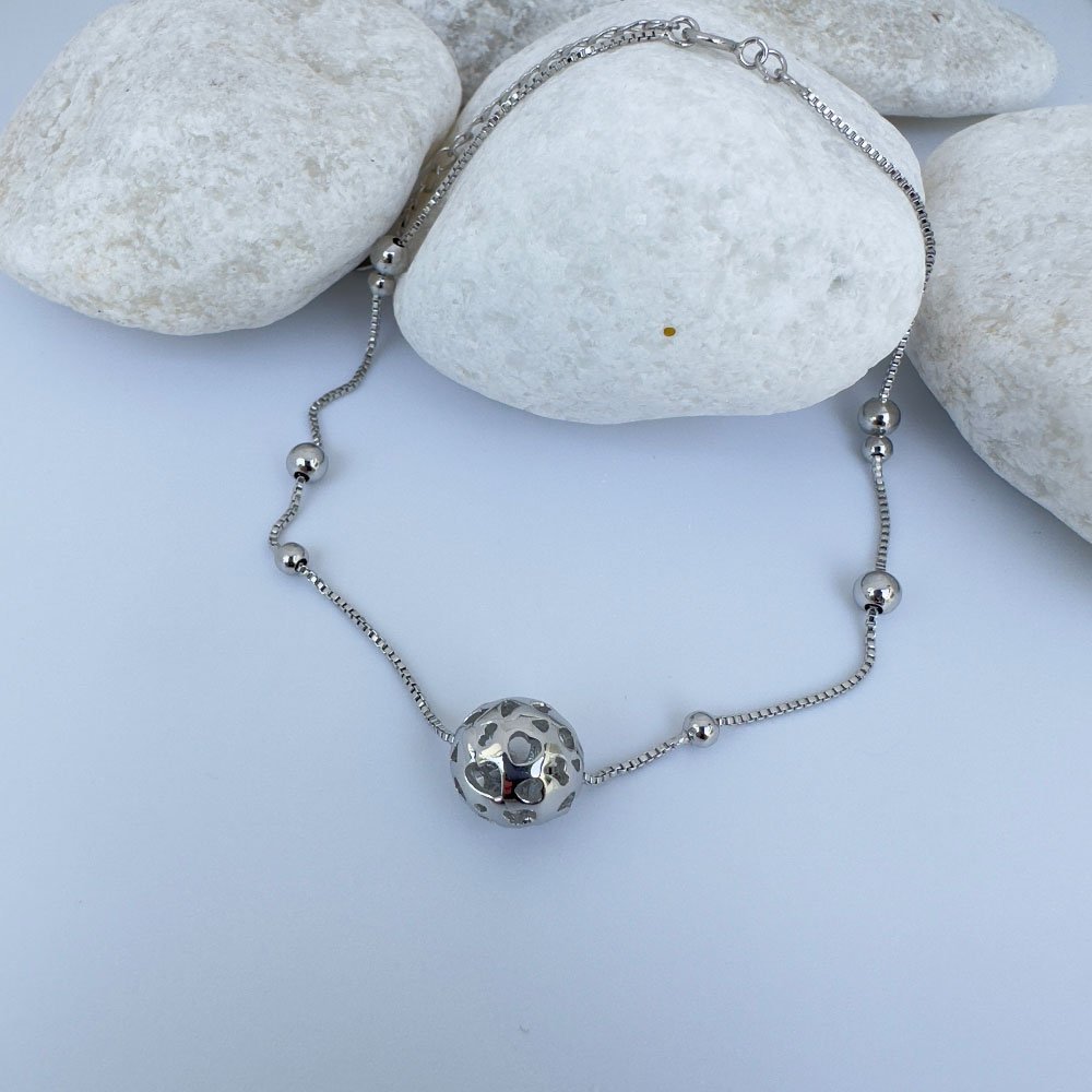 D8-Anklet-(2) Silver anklet with a sphere charm and smaller balls around the chain draped over white rocks.