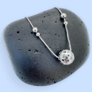Silver anklet with a large silver sphere and two smaller balls either side on a black rock with a blue background.