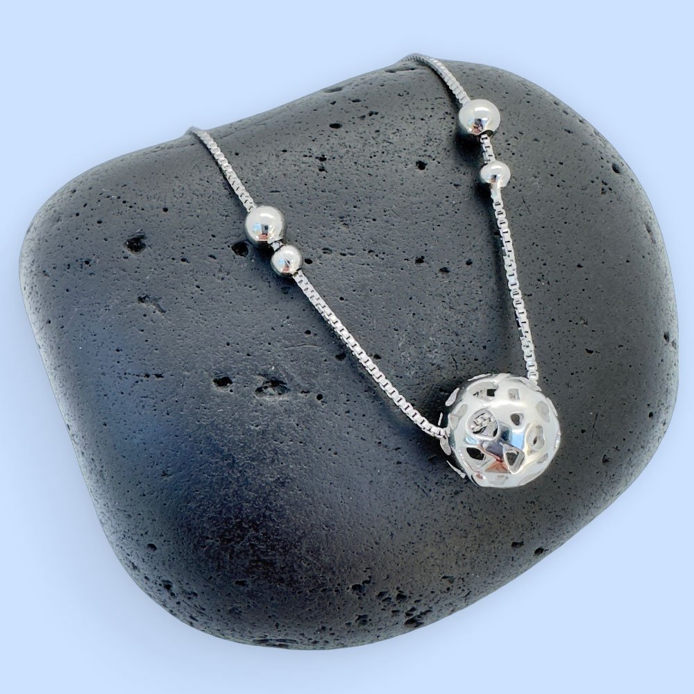D8-Anklet-rock Silver anklet with a large silver sphere and two smaller balls either side on a black rock with a blue background.