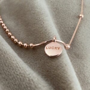 Rose gold anklet, one side with balls close together and another side with just a few far apart. There is a rose gold round pendant with the words lucky written on it in capital letters.