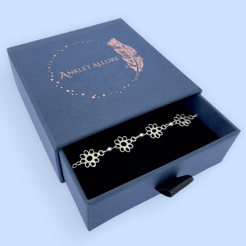 G13-Anklet-box Feminine silver anklets made up of flowers with eight petals linked together with a silver ball in between each flower, presented in a blue Anklet Allure jewellery box.