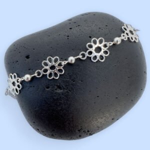 Feminine silver anklets made up of flowers with eight petals linked together with a silver ball in between each flower, presented on a black rock.