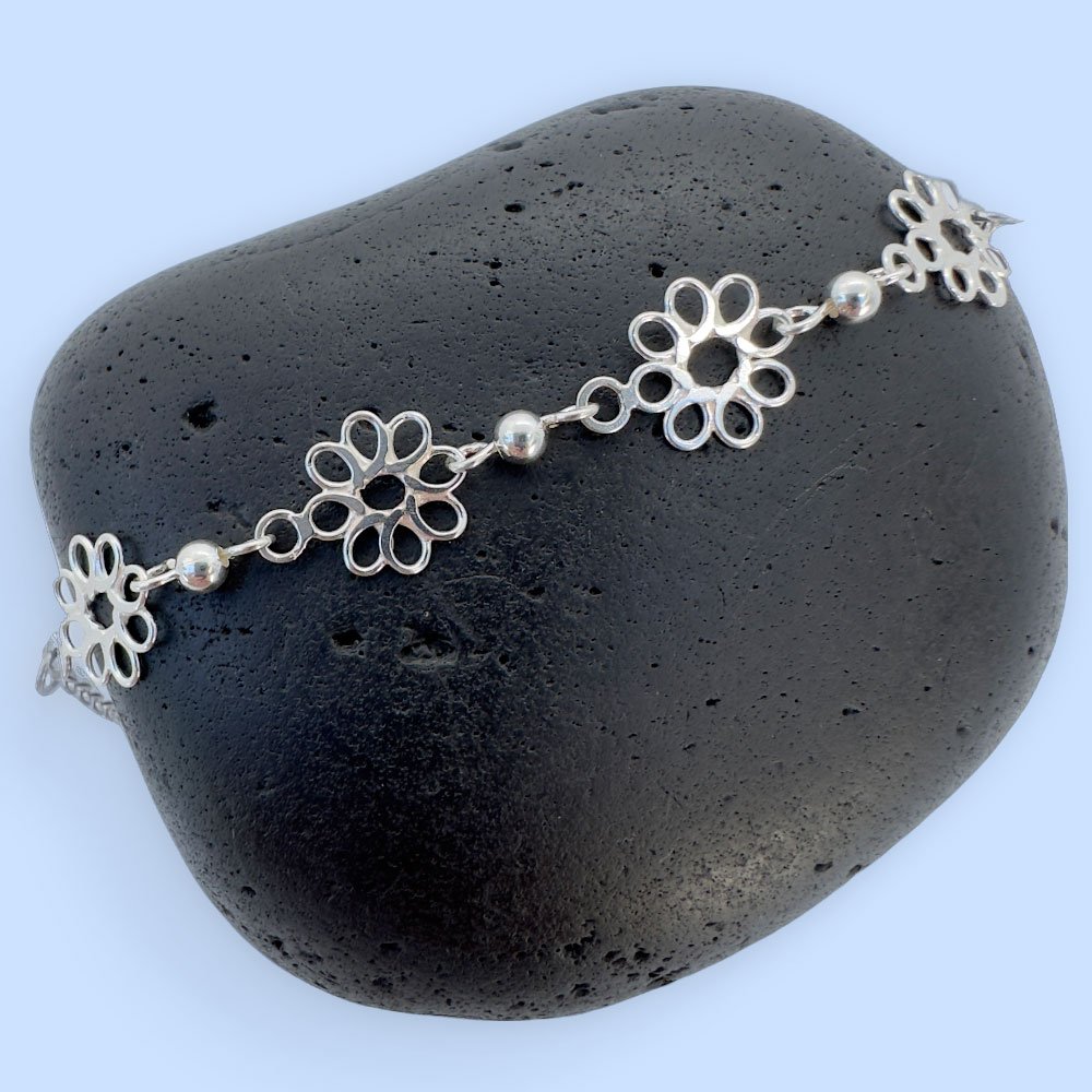 G13-Anklet-rock-(1) Feminine silver anklets made up of flowers with eight petals linked together with a silver ball in between each flower, presented on a black rock.
