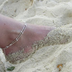 Close up of a sandy foot wearing a silver rhodium anklet.